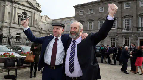 Getty Images Michael Healy Rae（左）与他的兄弟Danny Healy-Rae（右）。两人都穿着西装，手臂高举。Michael戴着帽子。